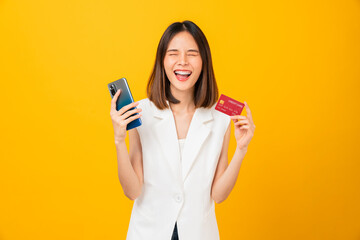 Cheerful young Asian woman holding smartphone with show mockup credit card on yellow background.