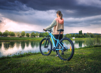 Woman riding a mountain bike near lake at overcast sunset in spring. Colorful landscape with sporty girl, bicycle, coast of river, green grass, cloudy sky in park in summer. Sport and travel. Biking