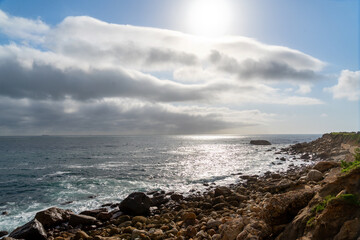 Fototapeta na wymiar coastal shoreline with distant horizon, rocky beach and sunshine through a clouded blue sky
