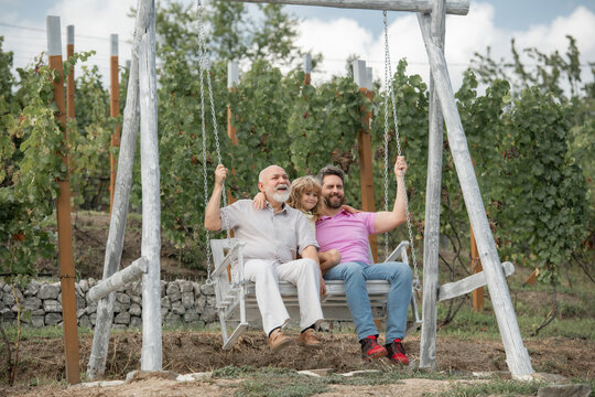 Boy son with father and grandfather swinging together in park outdoors. Three different generations ages grandfather father and child. Dad, son and granddad hugging.