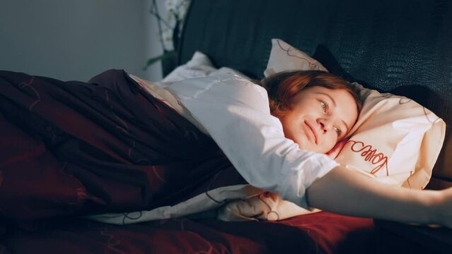 Smiling attractive sexy girl in white men's shirt goes to bed. Woman lies down on bed and puts head on pillow and covers herself with blanket. Female turns off lamp and closes eyes.