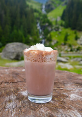 a glass of hot chocolate with cream on the background of a mountain waterfall