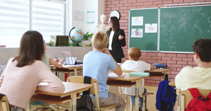 Cheerful Caucasian young female teacher of anatomy or biology talking to class and explaining human DNA to pupils. At school. Education concept. Indoors. Teaching. Lection about body.