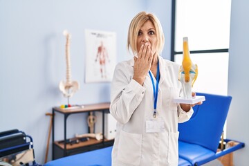 Middle age blonde traumatologist woman holding anatomical model of knee joint at clinic covering...