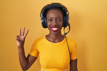 African woman with curly hair standing over yellow background wearing headphones showing and pointing up with fingers number three while smiling confident and happy.