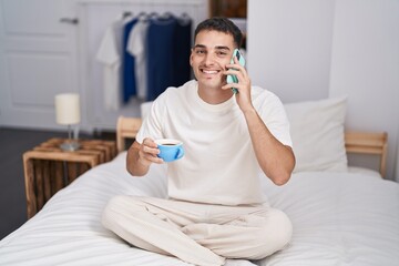 Young hispanic man talking on smartphone drinking coffee at bedroom