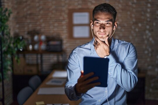 Handsome hispanic man working at the office at night looking confident at the camera smiling with crossed arms and hand raised on chin. thinking positive.