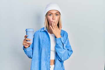 Beautiful blonde woman drinking a take away cup of coffee covering mouth with hand, shocked and...