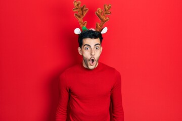 Young hispanic man wearing cute christmas reindeer horns afraid and shocked with surprise...