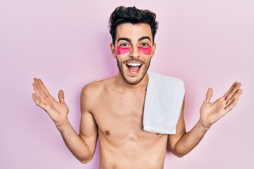 Young hispanic man shirtless wearing towel and eye bags patches celebrating victory with happy smile and winner expression with raised hands