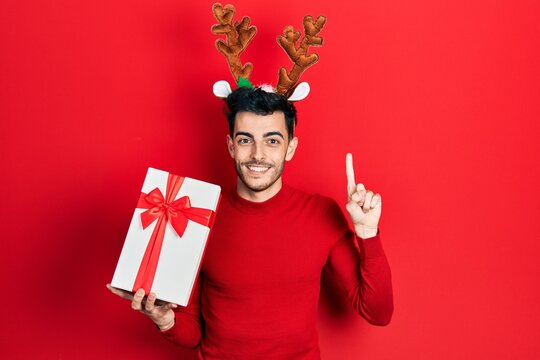 Young hispanic man wearing cute christmas reindeer horns holding gifts smiling with an idea or question pointing finger with happy face, number one