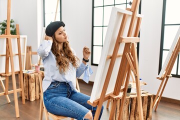 Young hispanic artist woman painting on canvas at art studio confuse and wondering about question....