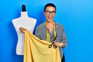 Beautiful hispanic woman with short hair standing by manikin holding cloth winking looking at the...