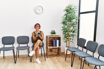 Young hispanic woman listening to music sitting on chair at waiting room