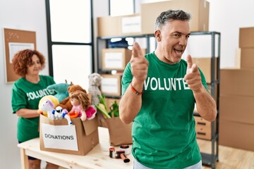 Middle age man wearing volunteer t shirt at donations stand pointing fingers to camera with happy and funny face. good energy and vibes.