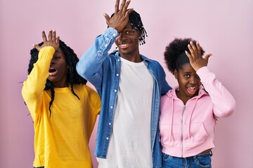 Group of three young black people standing together over pink background surprised with hand on...