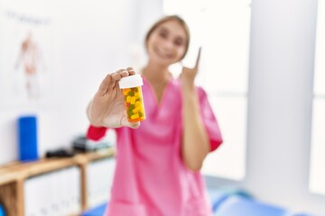 Young physiotherapist woman working at pain recovery clinic holding pills smiling with an idea or question pointing finger with happy face, number one