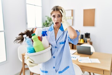 Young blonde woman wearing cleaner uniform holding cleaning products crazy and scared with hands on head, afraid and surprised of shock with open mouth
