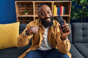 African american man holding television remote control pointing finger to one self smiling happy...