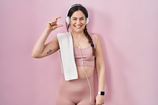Young brunette woman wearing sportswear and headphones smiling and confident gesturing with hand doing small size sign with fingers looking and the camera. measure concept.