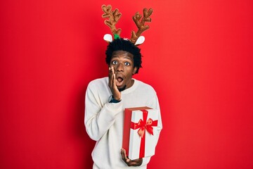 Young african american man wearing deer christmas hat holding gift afraid and shocked, surprise and...