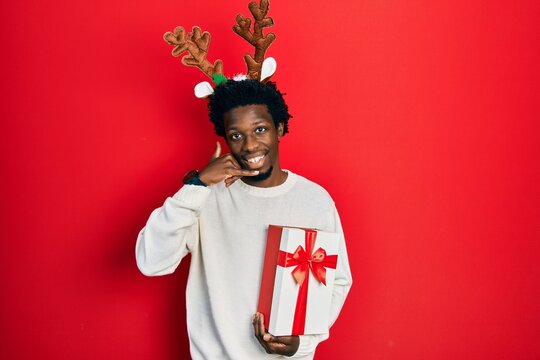 Young African American Man Wearing Deer Christmas Hat Holding Gift Smiling Doing Phone Gesture With Hand And Fingers Like Talking On The Telephone. Communicating Concepts.