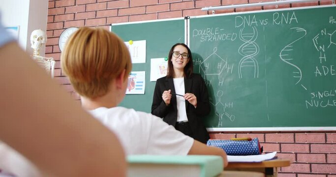 Caucasian young woman teacher of biology explaining DNA formulas and talking to pupils in classroom. Rear of kids at school listening to lesson. Educational concept. Discussion. Question answering.