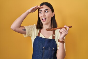 Hispanic girl eating healthy  wooden spoon very happy and smiling looking far away with hand over head. searching concept.