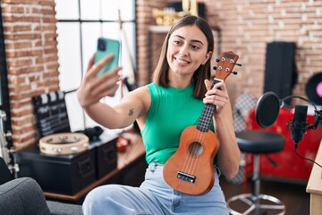 Fototapeta na wymiar Young hispanic woman musician make selfie by the smartphone holding ukelele at music studio