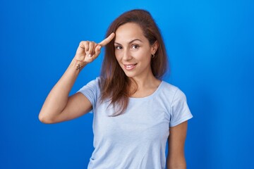 Brunette woman standing over blue background smiling pointing to head with one finger, great idea or thought, good memory