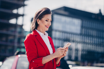 Photo of cheerful positive mature lady wear red jacket walking chatting modern device outside city...