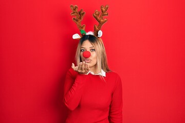 Beautiful hispanic woman wearing deer christmas hat and red nose looking at the camera blowing a...