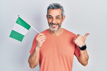 Handsome middle age man with grey hair holding nigeria flag pointing thumb up to the side smiling...