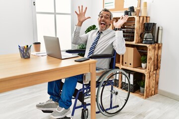 Middle age hispanic man working at the office sitting on wheelchair celebrating crazy and amazed for success with arms raised and open eyes screaming excited. winner concept