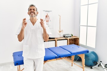 Middle age hispanic therapist man working at pain recovery clinic gesturing finger crossed smiling with hope and eyes closed. luck and superstitious concept.