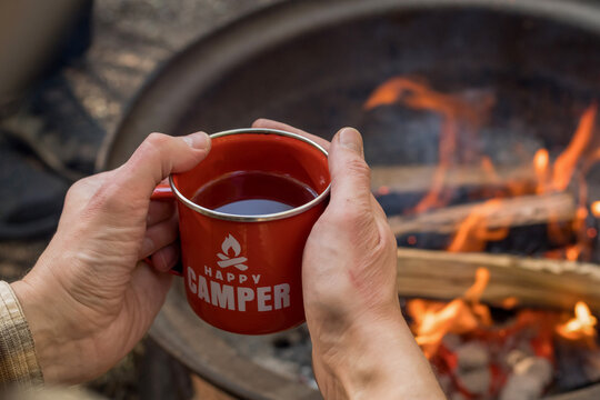 Camping lifestyle concept. Hands holding red enamel mug near a campfire. Happy camper