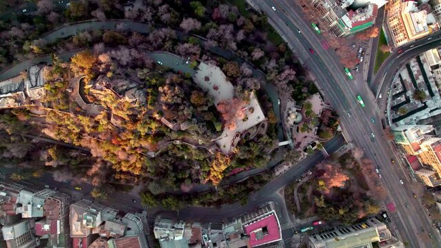 Aerial top down dolly in of Santa Lucia Hill with autumnal trees, traffic in Alameda avenue and Santiago buildinds at sunset, Chile