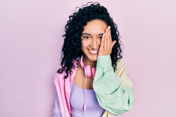 Young hispanic woman with curly hair wearing gym clothes and using headphones covering one eye with hand, confident smile on face and surprise emotion.