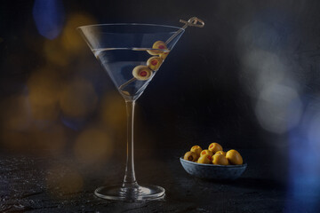 Martini, a glass with spicy olives, on a black background. Alcoholic drink with bokeh, toned image