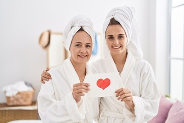Middle age woman and daughter wearing bath robe holding heart card smiling with a happy and cool...