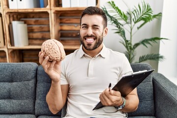 Handsome hispanic man holding brain working at psychology clinic sticking tongue out happy with...