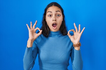 Young brazilian woman standing over blue isolated background looking surprised and shocked doing ok approval symbol with fingers. crazy expression