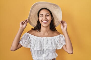 Young brazilian woman wearing summer hat over yellow background smiling and laughing hard out loud because funny crazy joke.