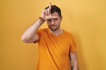 Fototapeta na wymiar Young hispanic man standing over yellow background making fun of people with fingers on forehead doing loser gesture mocking and insulting.