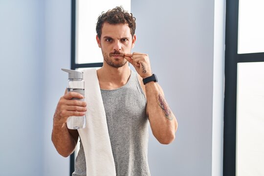 Young hispanic man wearing sportswear drinking water mouth and lips shut as zip with fingers. secret and silent, taboo talking