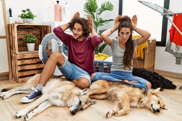 Young hispanic couple doing laundry with dogs doing bunny ears gesture with hands palms looking...