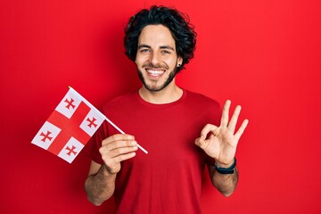Handsome hispanic man holding georgia flag doing ok sign with fingers, smiling friendly gesturing excellent symbol