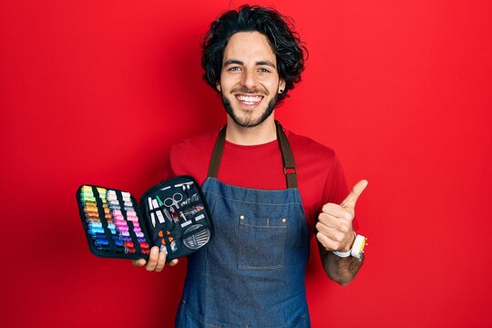 Handsome hispanic man wearing apron holding sew kit smiling happy and positive, thumb up doing excellent and approval sign