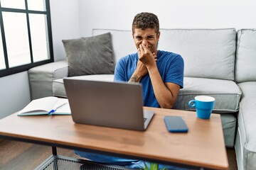 Young handsome hispanic man using laptop sitting on the floor smelling something stinky and disgusting, intolerable smell, holding breath with fingers on nose. bad smell