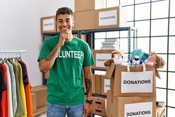 Young handsome hispanic man wearing volunteer t shirt at donations stand with hand on chin thinking about question, pensive expression. smiling and thoughtful face. doubt concept.